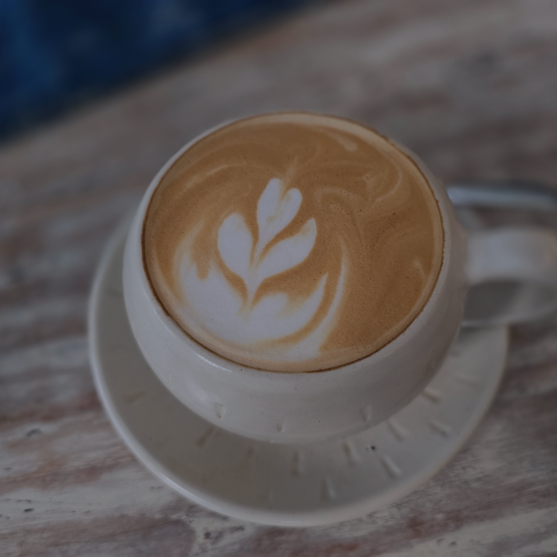 A cup of latte coffee with leaf-shaped latte art, served in a white ceramic cup and saucer on a rustic wooden table. The Mayne Bean on Mayne Island
