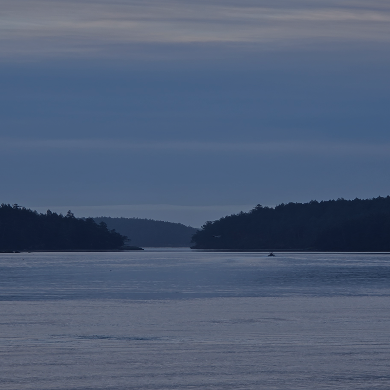 A calm blue lake or river with dark tree-lined islands on both sides under a muted blue-gray sky. Mayne Island. 