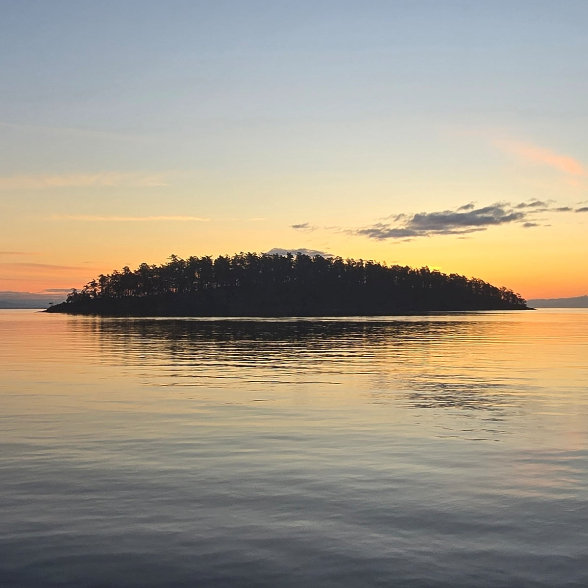 A tranquil sunset seascape with a small tree-covered island silhouetted against a warm orange and pale blue sky, reflected in calm water.